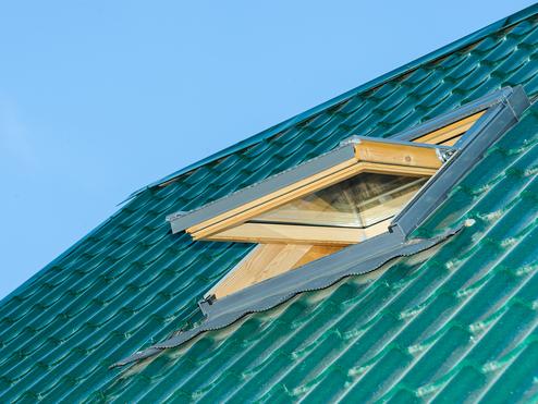 Dachfläche mit grünem Ziegeldach und geöffnetem Dachfenster unter klarem Himmel.